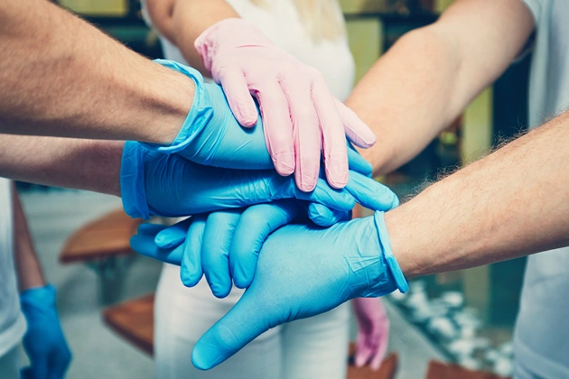 Team members stack hands together in unity gesture, wearing blue gloves indoors.
