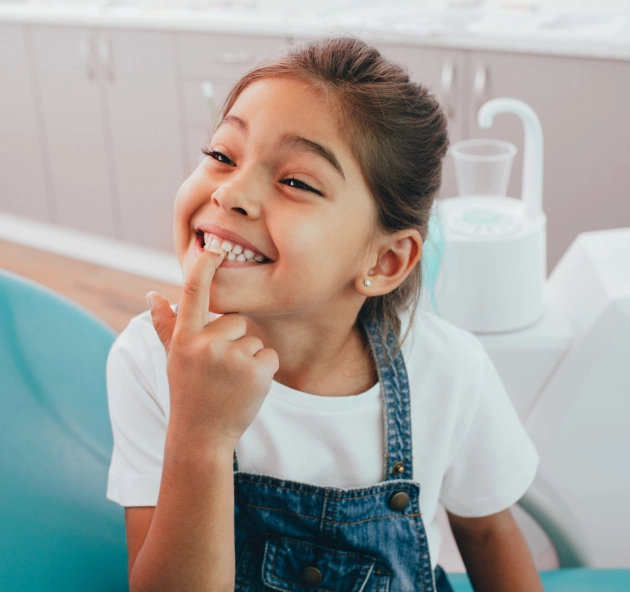 Young girl smiling in dental office, touching her teeth with finger, wearing apron.