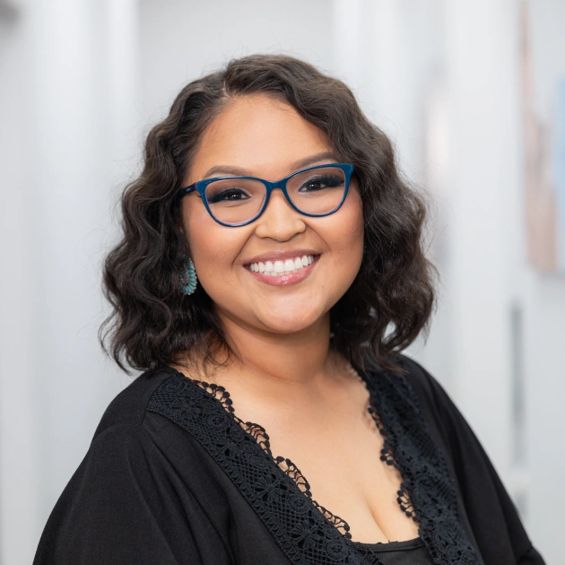Professional headshot of smiling woman wearing glasses and black top against blurred office background.