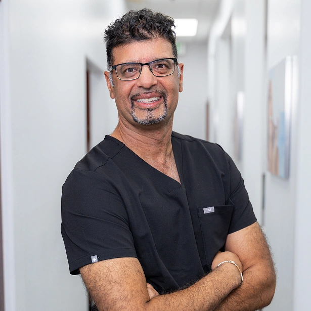 Male dentist in black scrubs smiling in modern dental office hallway.