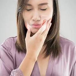 Woman touching her cheek in pain, wearing purple shirt against white background.