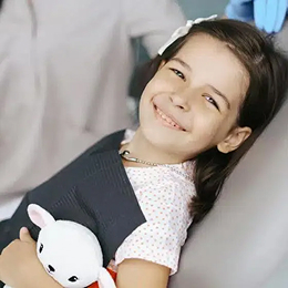 Young girl smiling at dental office, holding stuffed animal, wearing dark jacket.