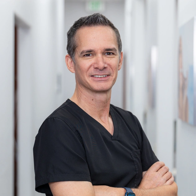 Male dental professional in black scrubs smiling with crossed arms in clinical hallway.