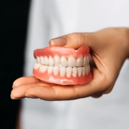 Hand holding full denture model showing white teeth against neutral background.