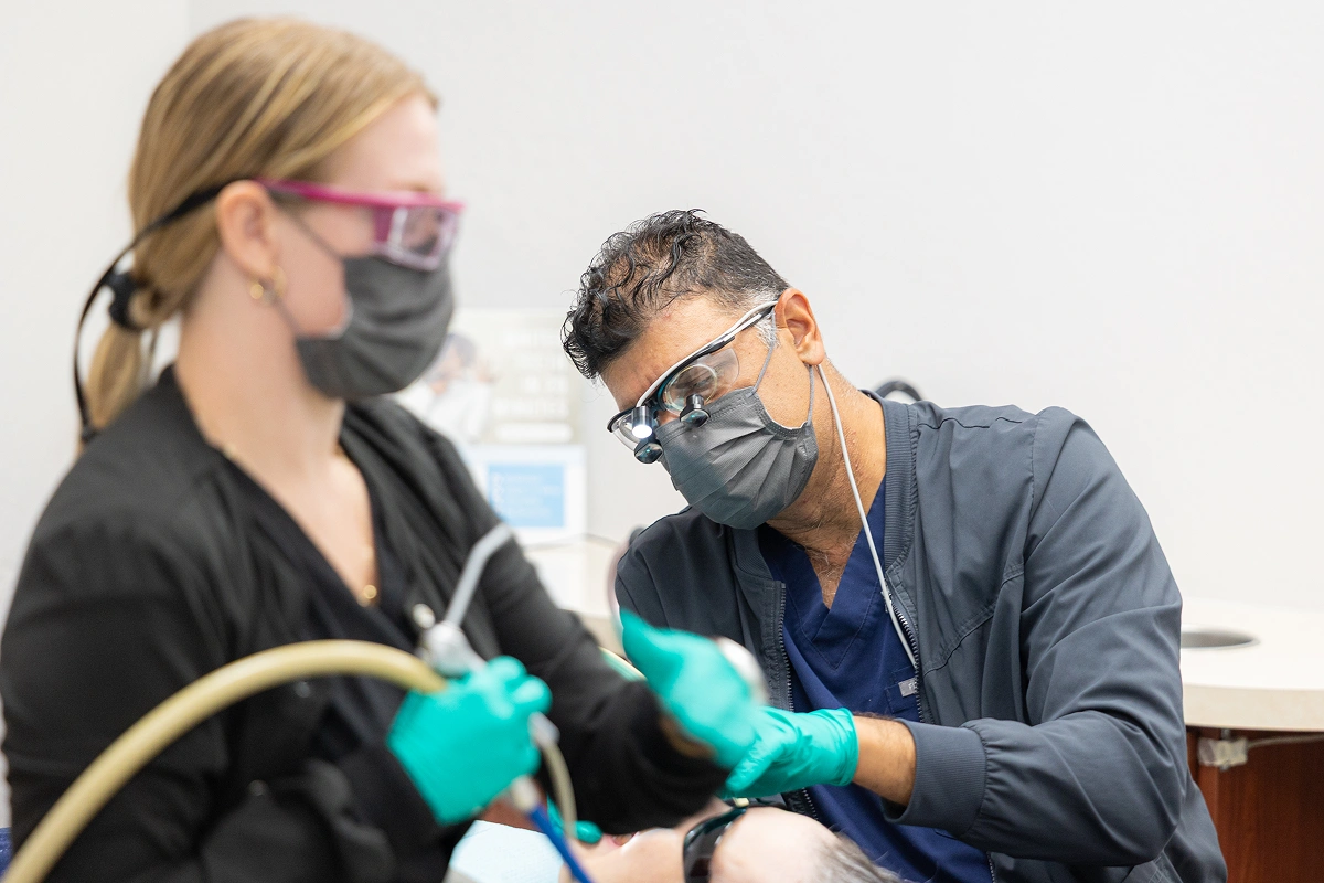 Two dental professionals wearing masks and gloves examine a dental model in a clinical setting.
